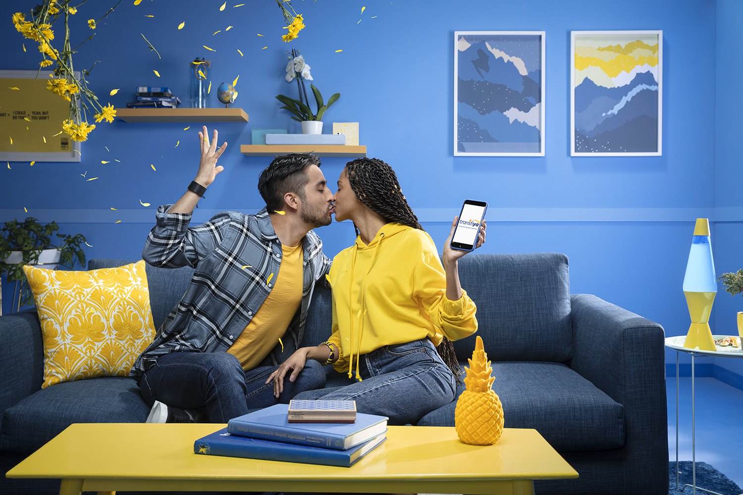 A couple on a blue sofa in a blue room, yellow cushions and a phone between them
