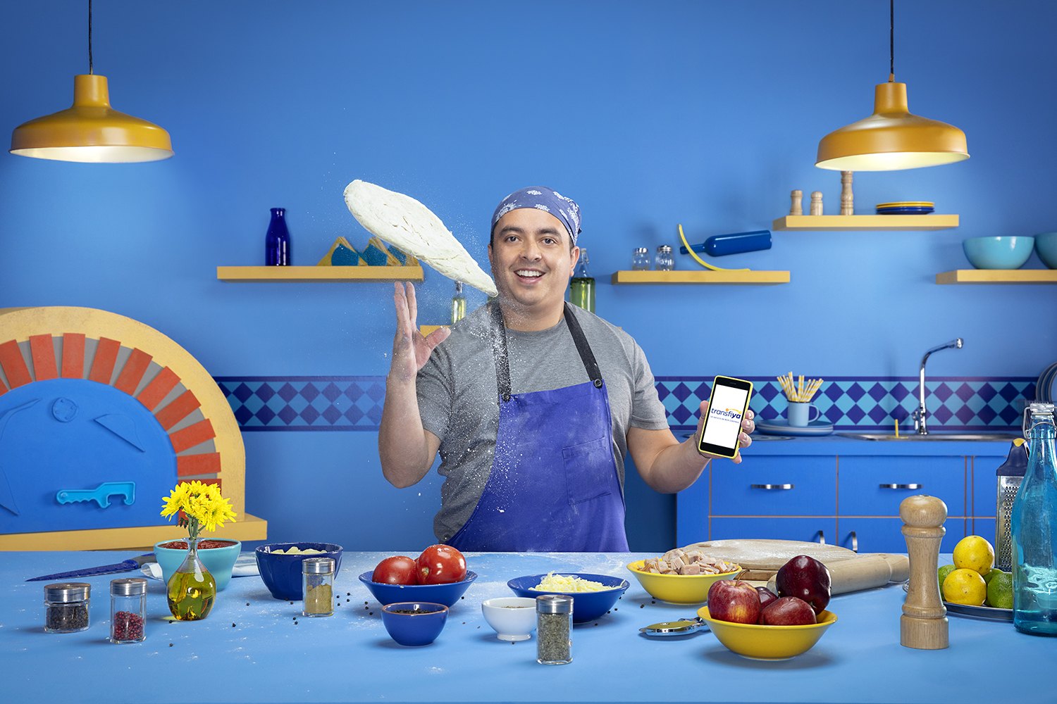 A man tossing dough in a blue kitchen hung with yellow pendant lamps