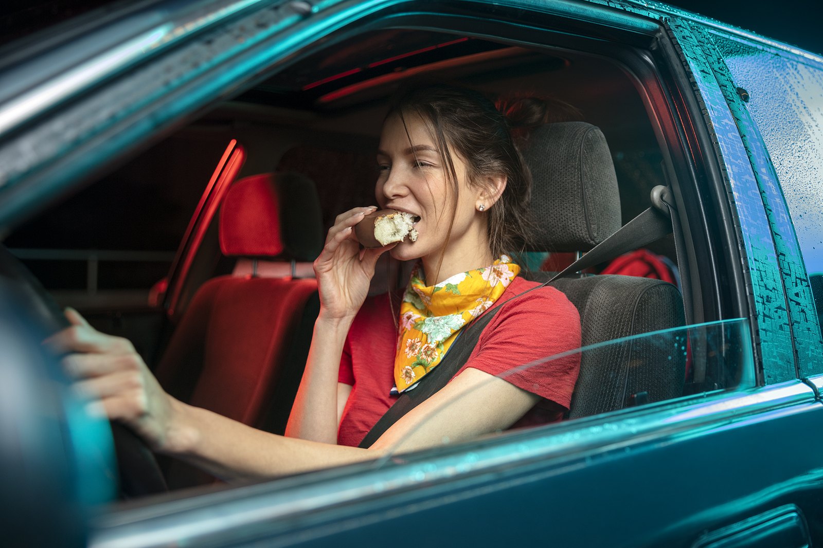 A woman at the wheel of a parked car takes an enormous bite, streetlight raking across the windscreen