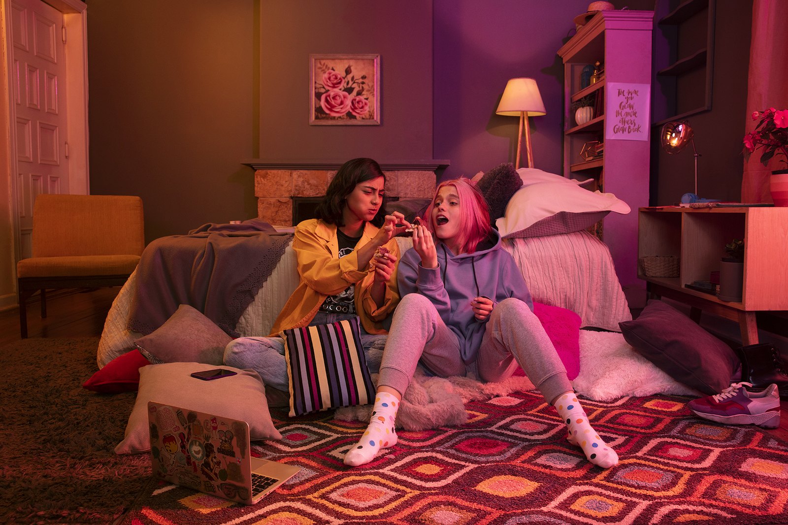 Two friends sitting on a bedroom floor mid-laugh, the whole room washed in pink and magenta