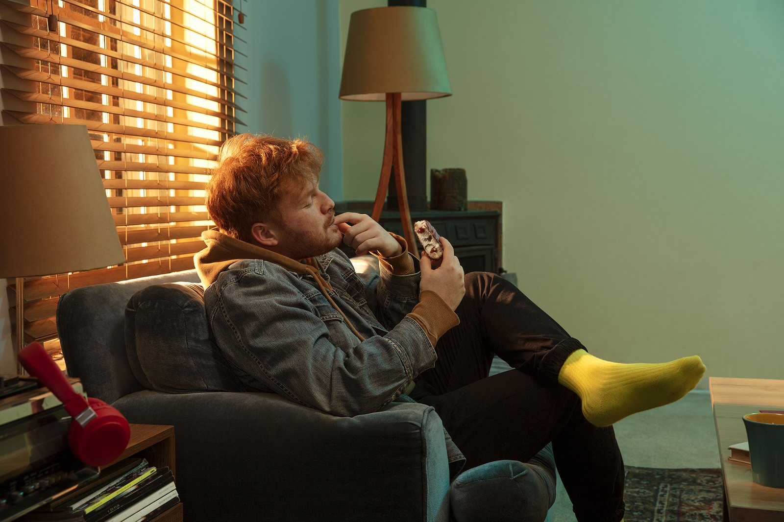 A man sunk deep into a sofa eating in green light, one yellow sock up on the table