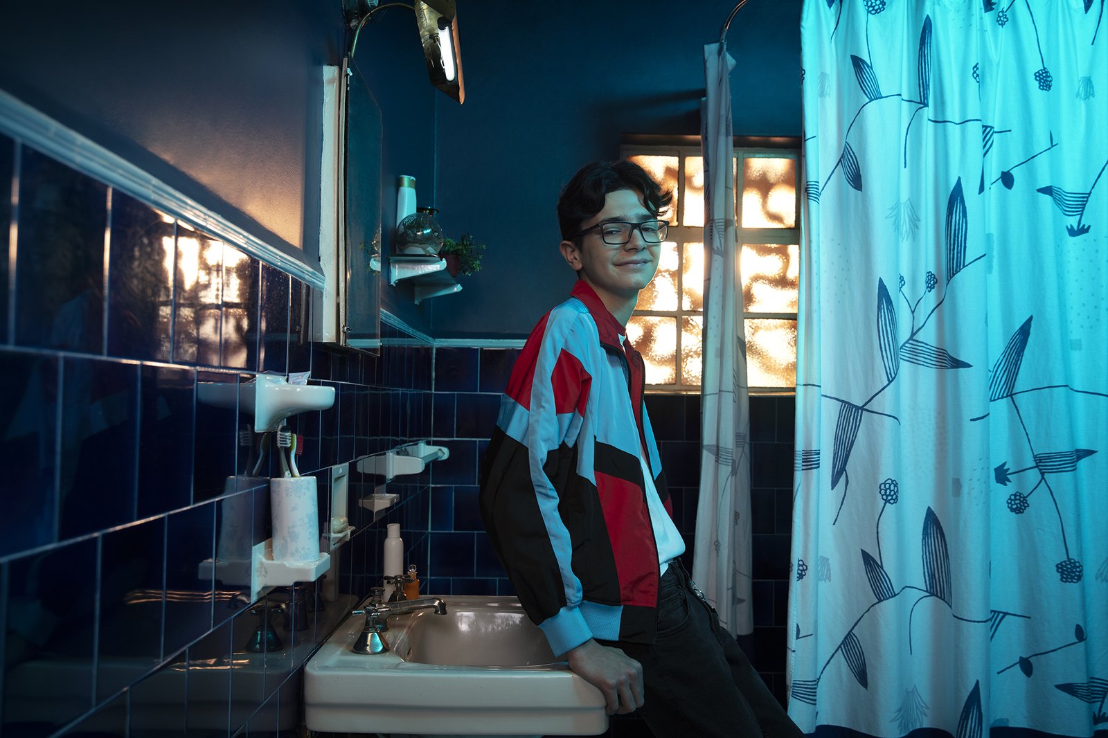 A figure leaning against the sink of a blue-tiled bathroom, patterned shower curtain glowing beside him