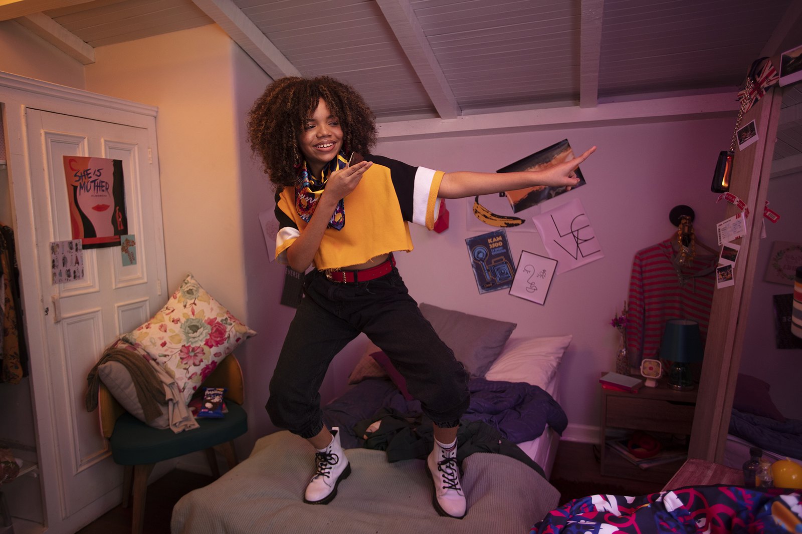 A woman standing on her bed in a pink attic room, arm flung out, clothes everywhere
