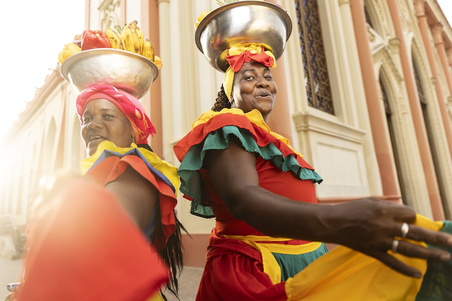 Two palenqueras in profile against yellow colonial walls, bowls held steady