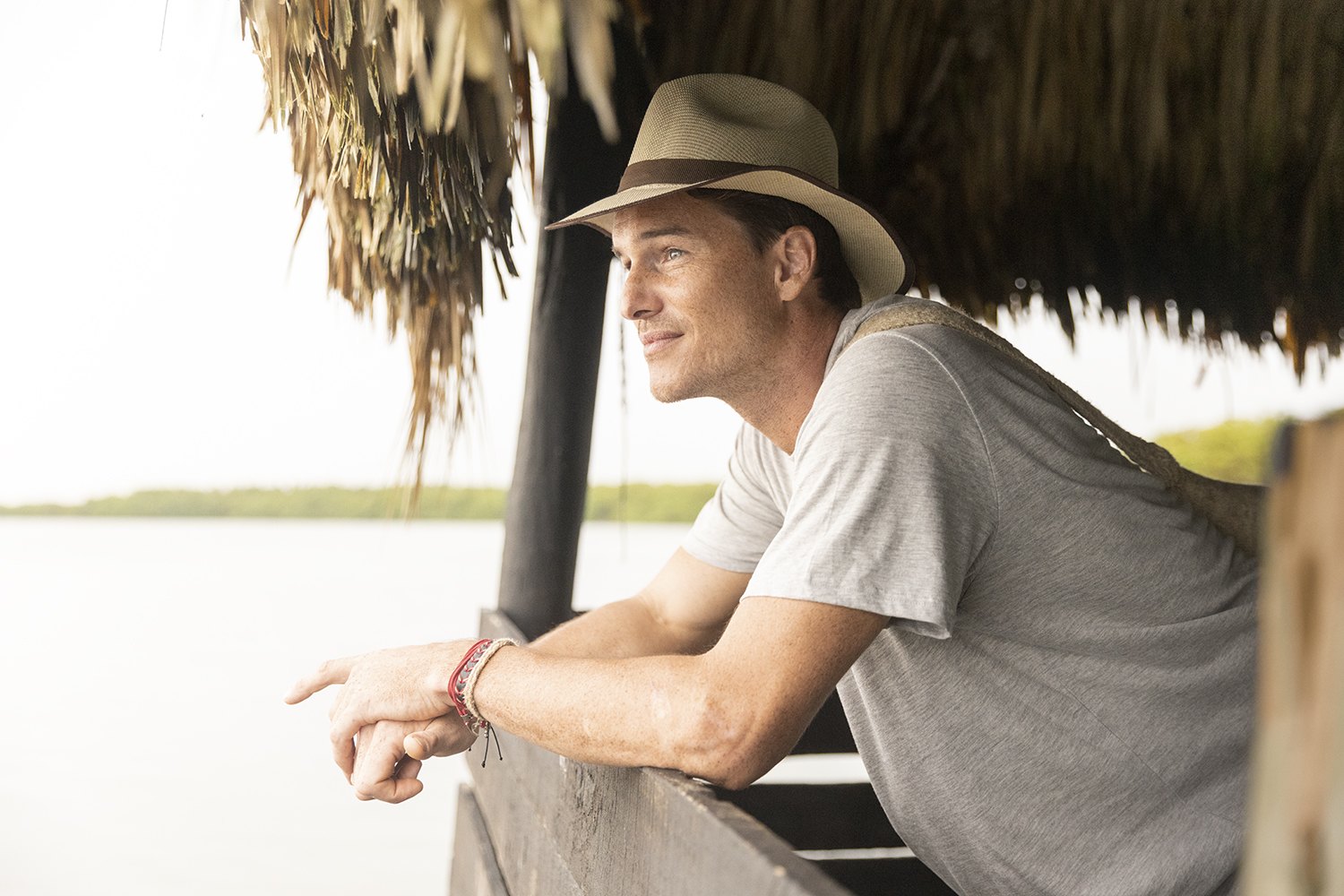 A traveller leaning on a rail beneath a palm-thatch roof looking out at the water