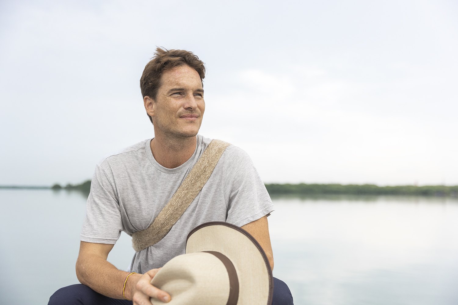 A man seated by the shoreline turning a straw hat in his hands