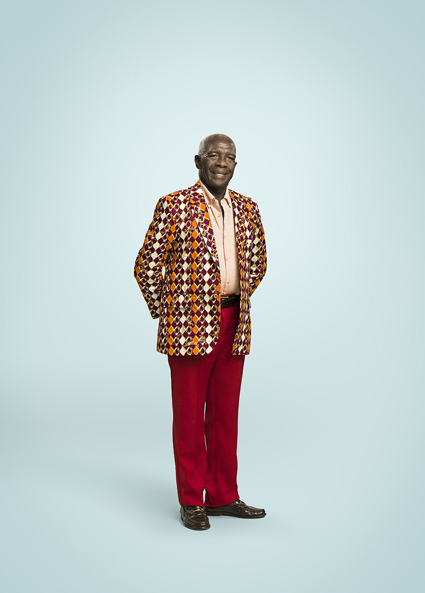 A man in a patterned jacket and red trousers standing full length on pale blue