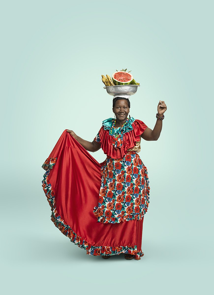 A dancer in a red ruffled dress swings her skirt, fruit balanced on her head