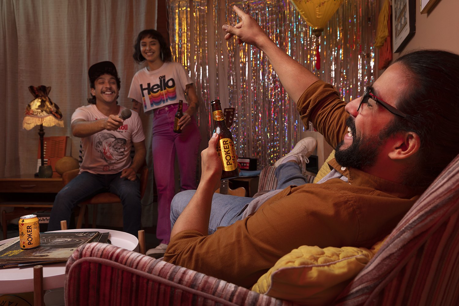 A man sunk into an armchair holding a bottle while friends celebrate behind him under a gold tinsel curtain