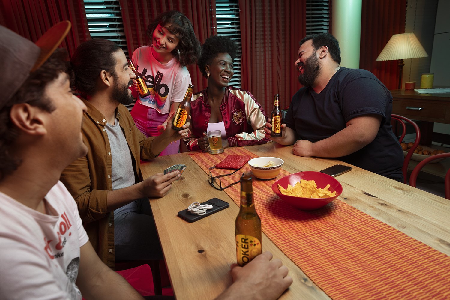 Five friends around a wooden table mid-card-game, bottles and a bowl of crisps between them