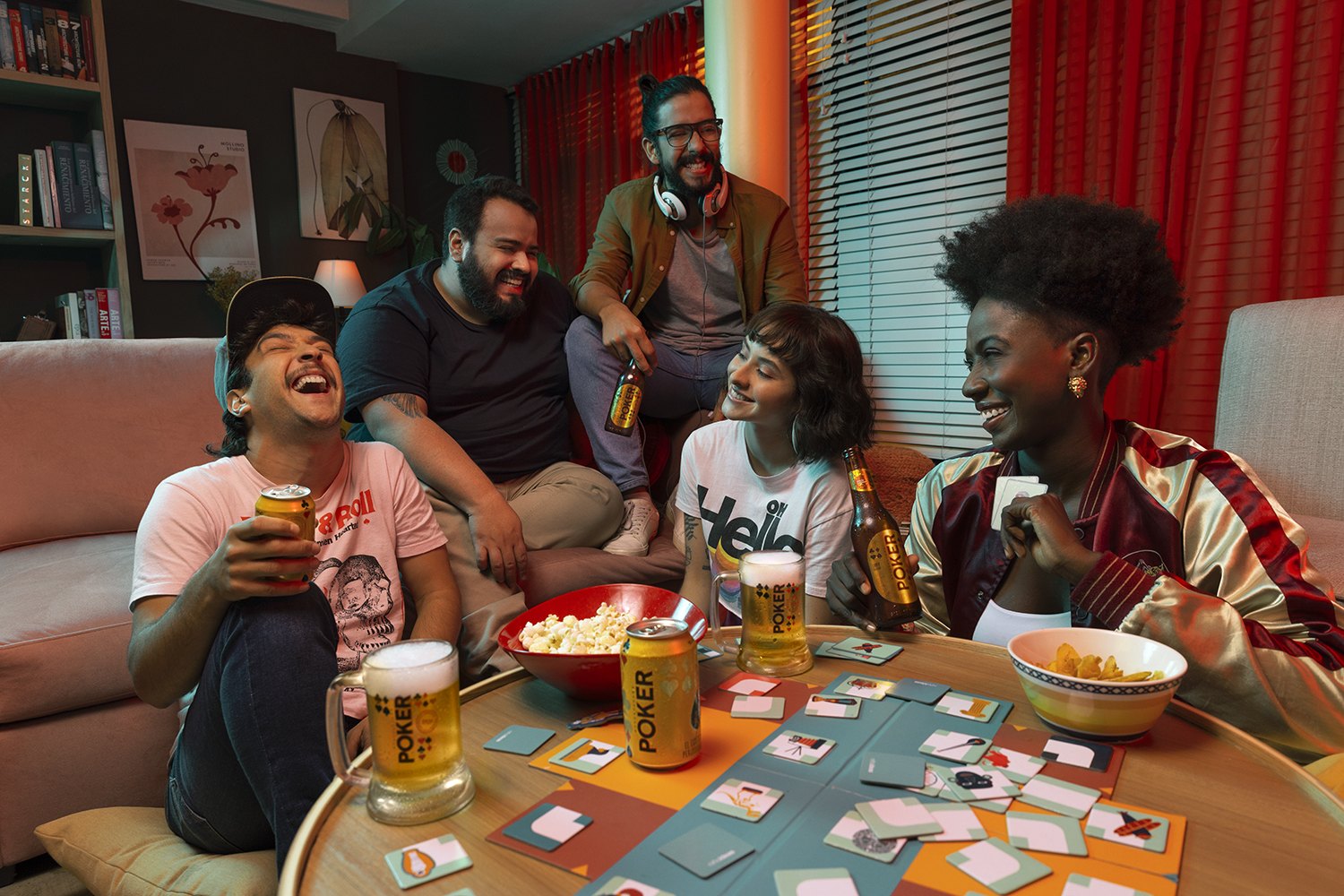 The full group around a low table covered in cards, glasses and snacks, red curtains behind