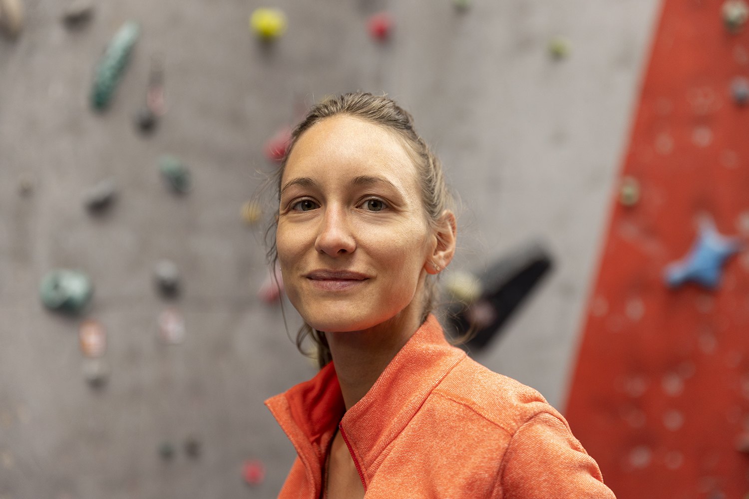 A climber in an orange top smiling, the holds of the wall soft behind her
