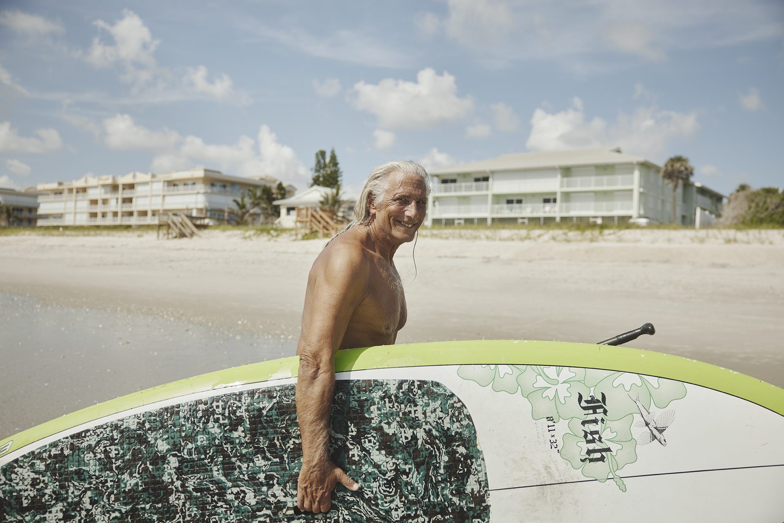 An older surfer smiles beside his board on the sand