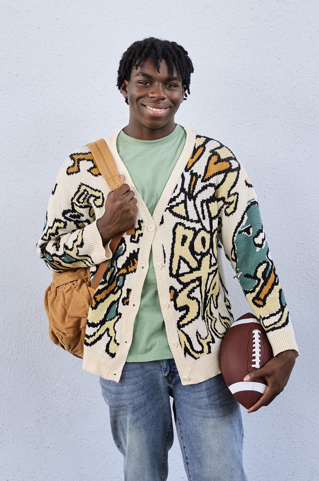 A student in a patterned cardigan holding a football, grey seamless behind