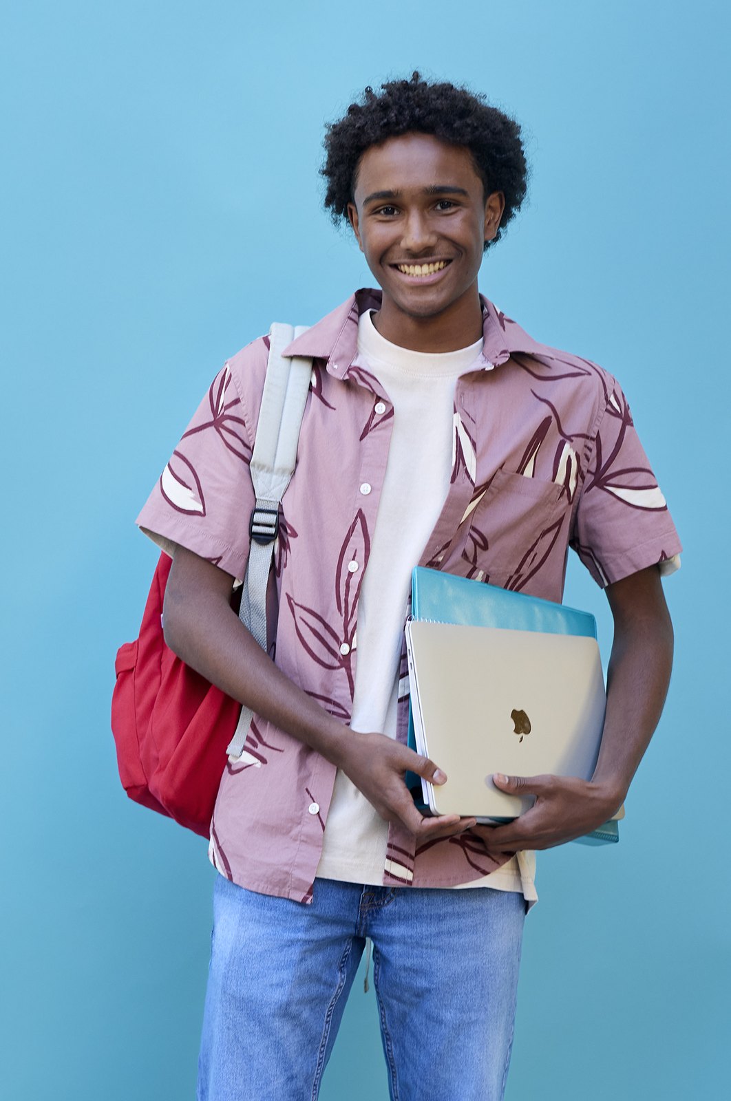 A student in a patterned shirt with a red bag and a laptop under one arm