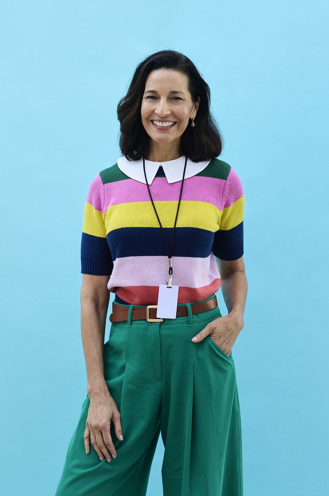 An educator in a striped polo with a lanyard, laptop tucked under the arm, on a teal backdrop