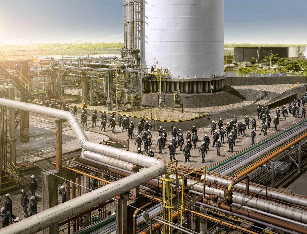 Refinery pipework in the foreground with a mass of workers gathered beyond