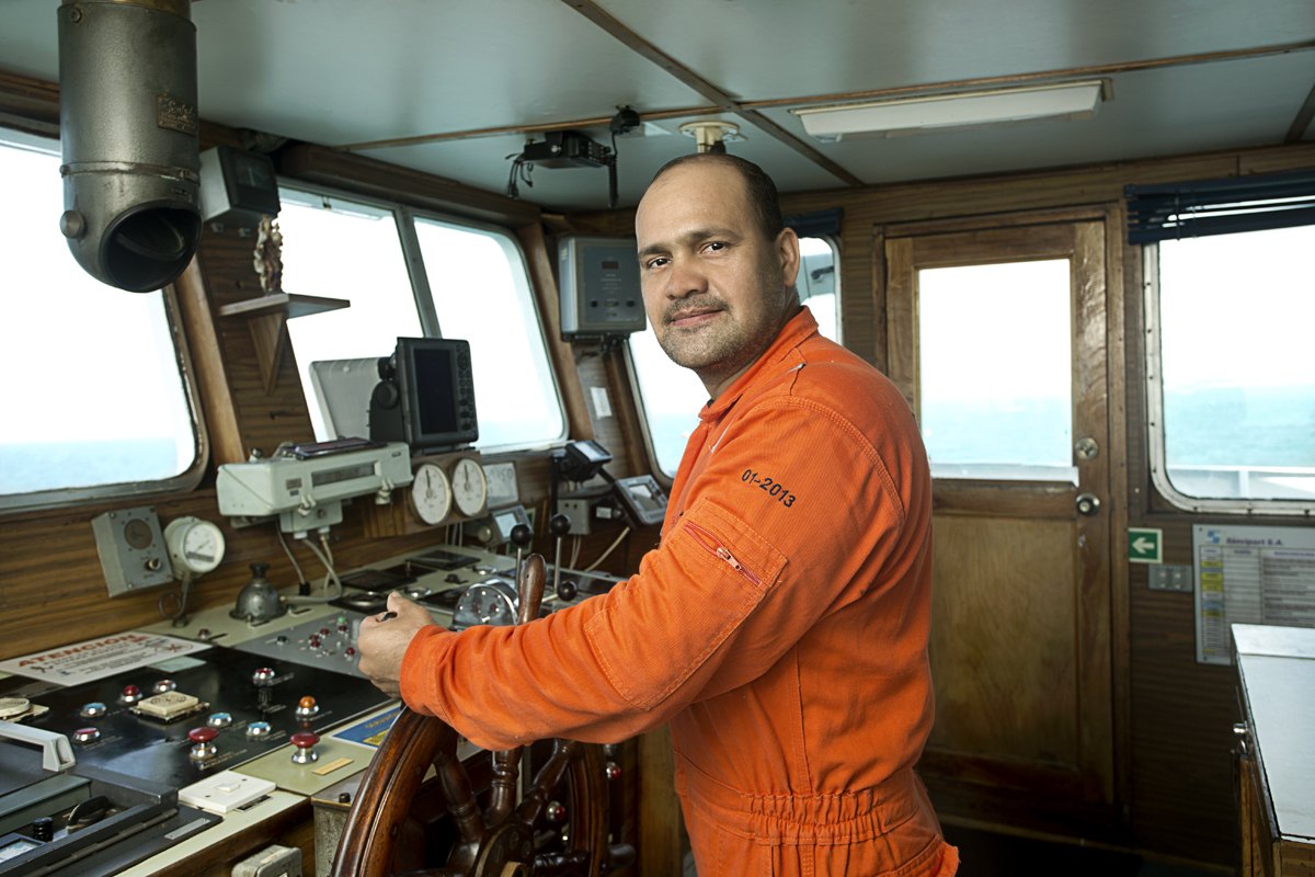An engineer in orange coveralls at the controls inside a vessel wheelhouse