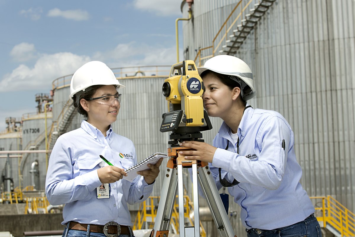 Two surveyors setting a theodolite with storage tanks rising behind them