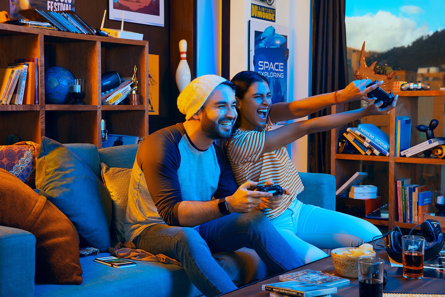 Two friends on a sofa mid-game, controllers up, shelves of books behind
