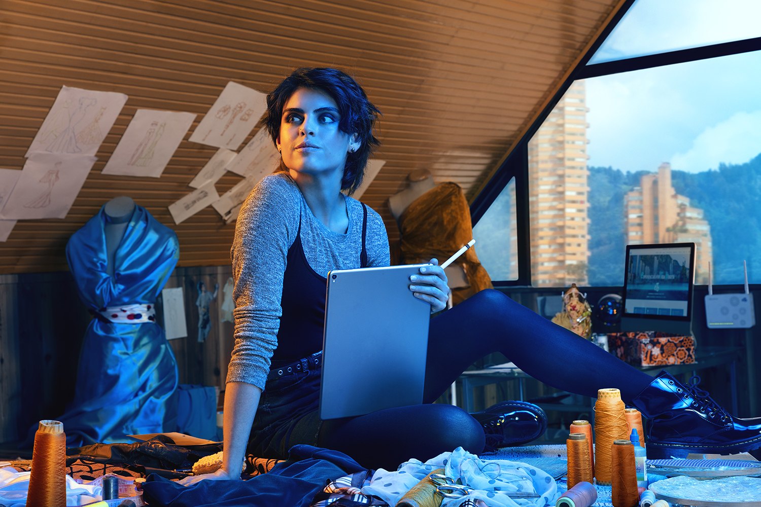 A woman working on a tablet on an unmade bed, city rooftops through the window
