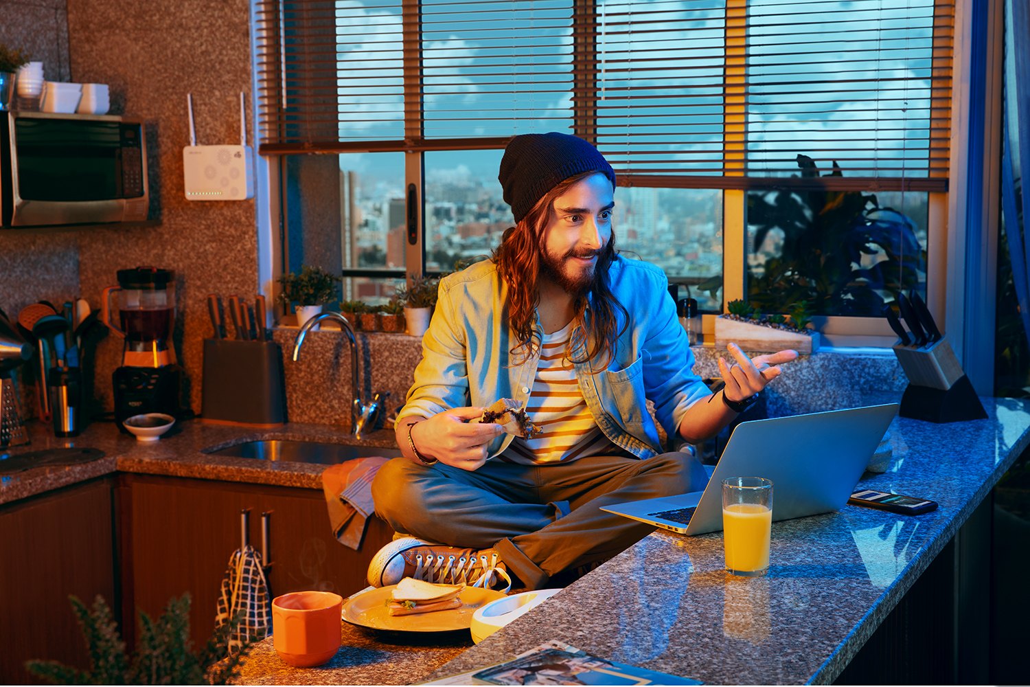 A man at a kitchen counter with a laptop and breakfast, window light behind