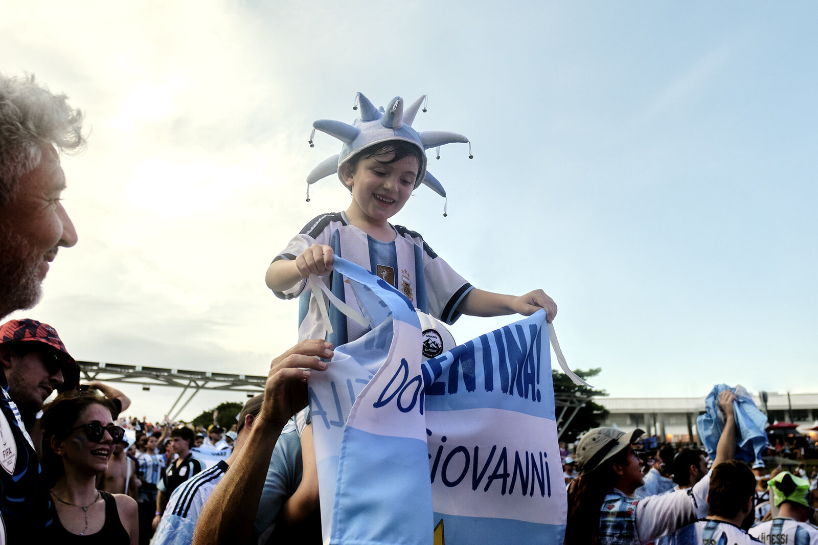 A young Argentina fan in a jester hat rides above the crowd with a hand-lettered banner