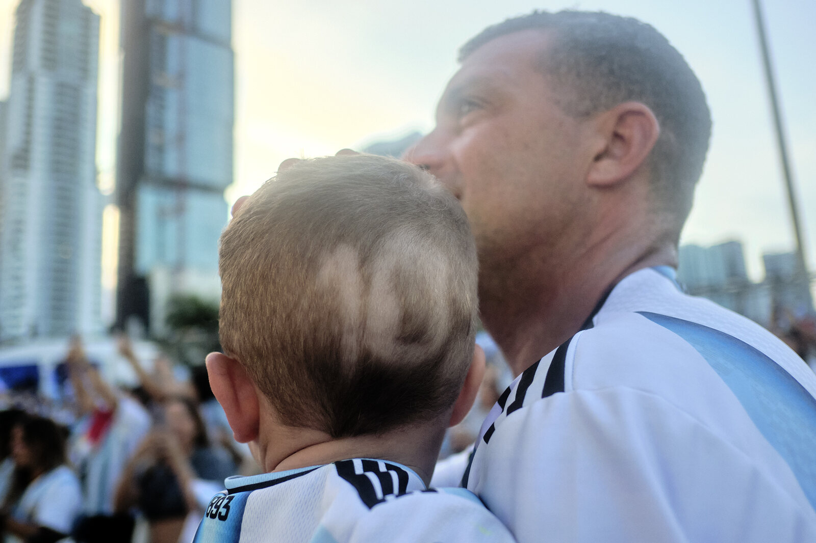 A father in an Argentina shirt holds his son close, both watching the screen