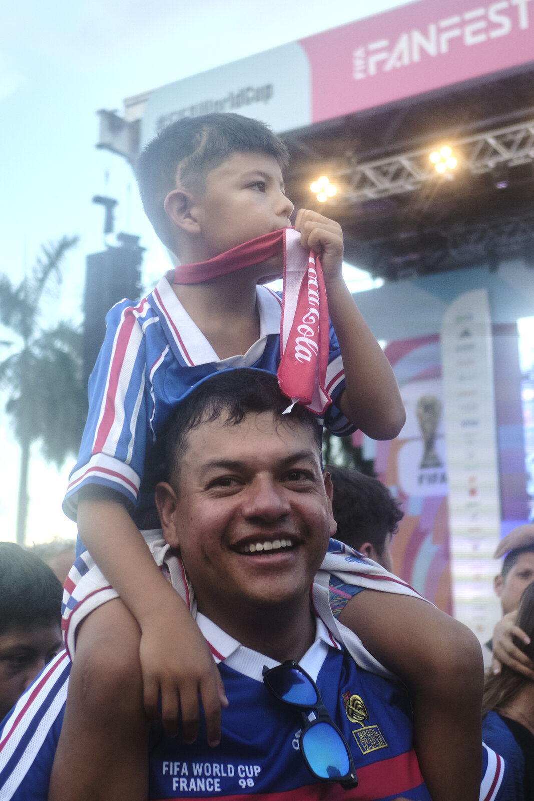 A boy rides his father's shoulders, both in vintage France World Cup jerseys