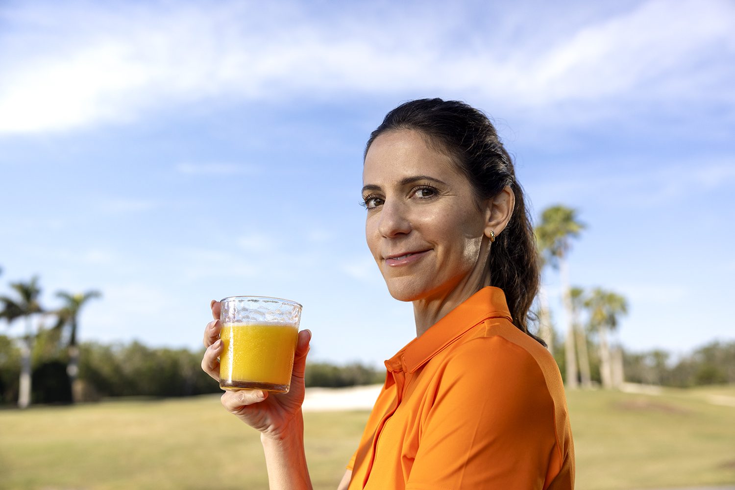 A woman in orange holds a glass of fresh orange juice on a golf course