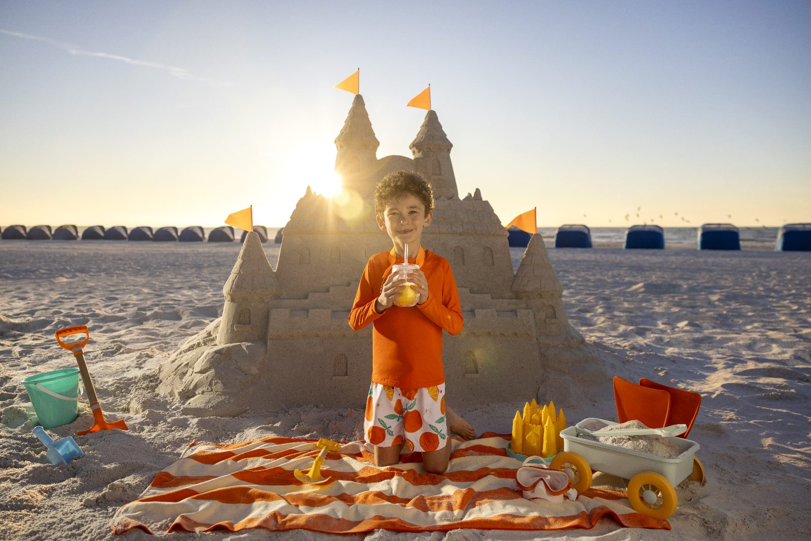 A child builds a sandcastle on the beach with orange juice nearby