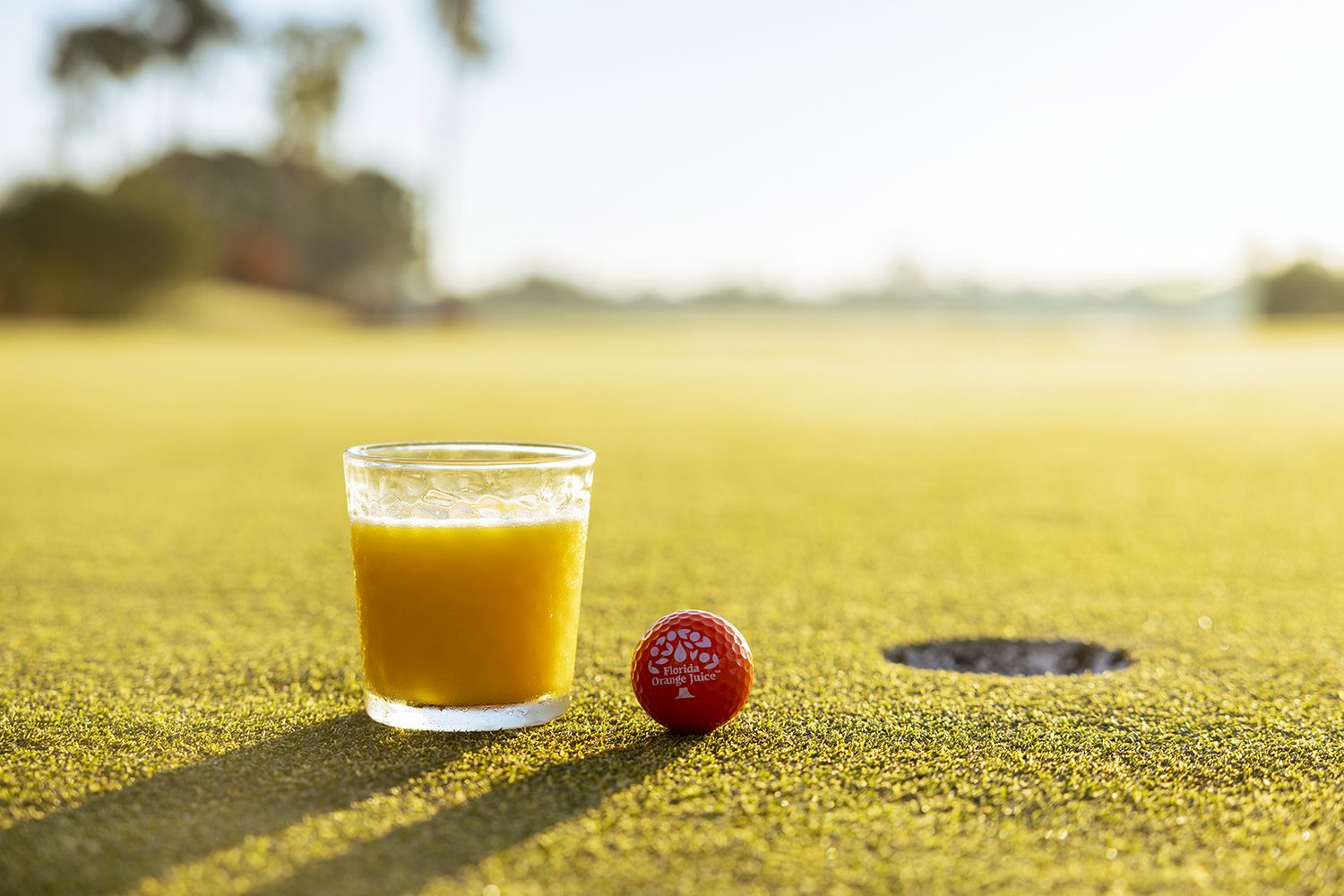 A glass of orange juice rests on the green beside a golf hole