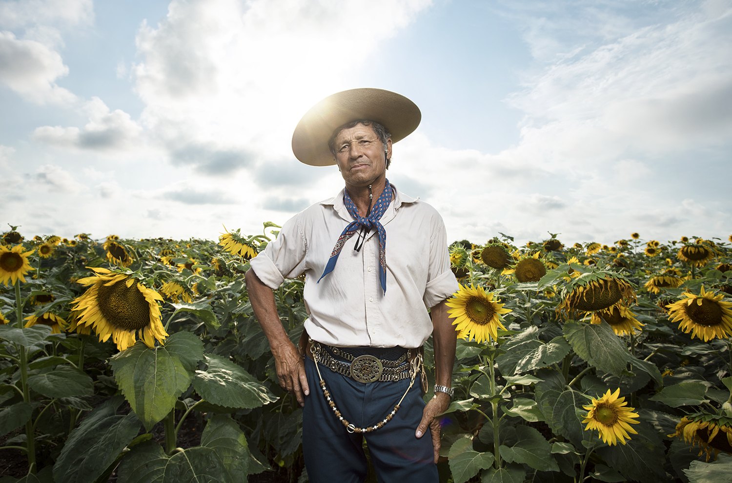 A gaucho in a wide-brimmed hat and neckerchief stands chest-deep in a sunflower field with the sun behind him