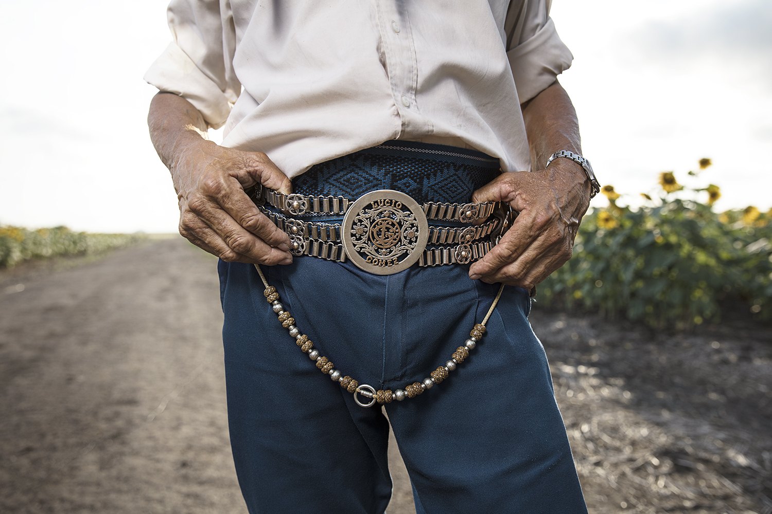 Close detail of weathered hands resting on an ornate silver rastra belt over dark bombachas