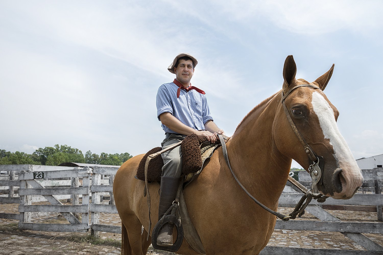 A young rider in a red neckerchief sits a chestnut horse beside the white corral rails