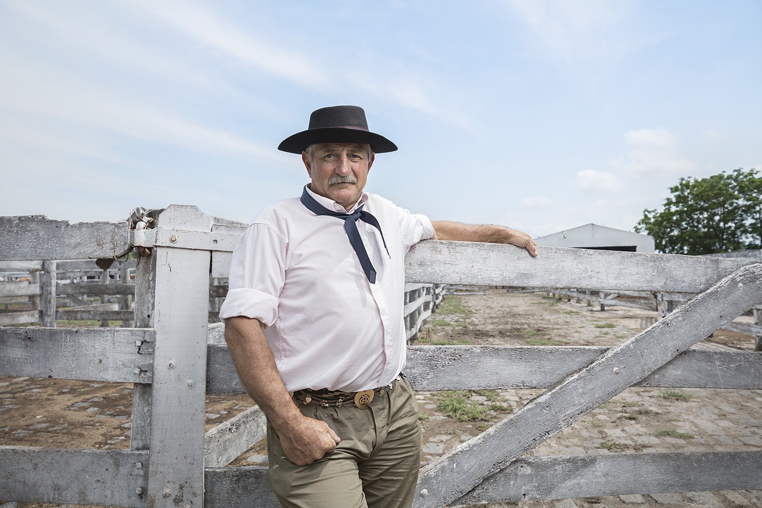 An older gaucho in a black hat and blue neckerchief leans against a whitewashed corral gate
