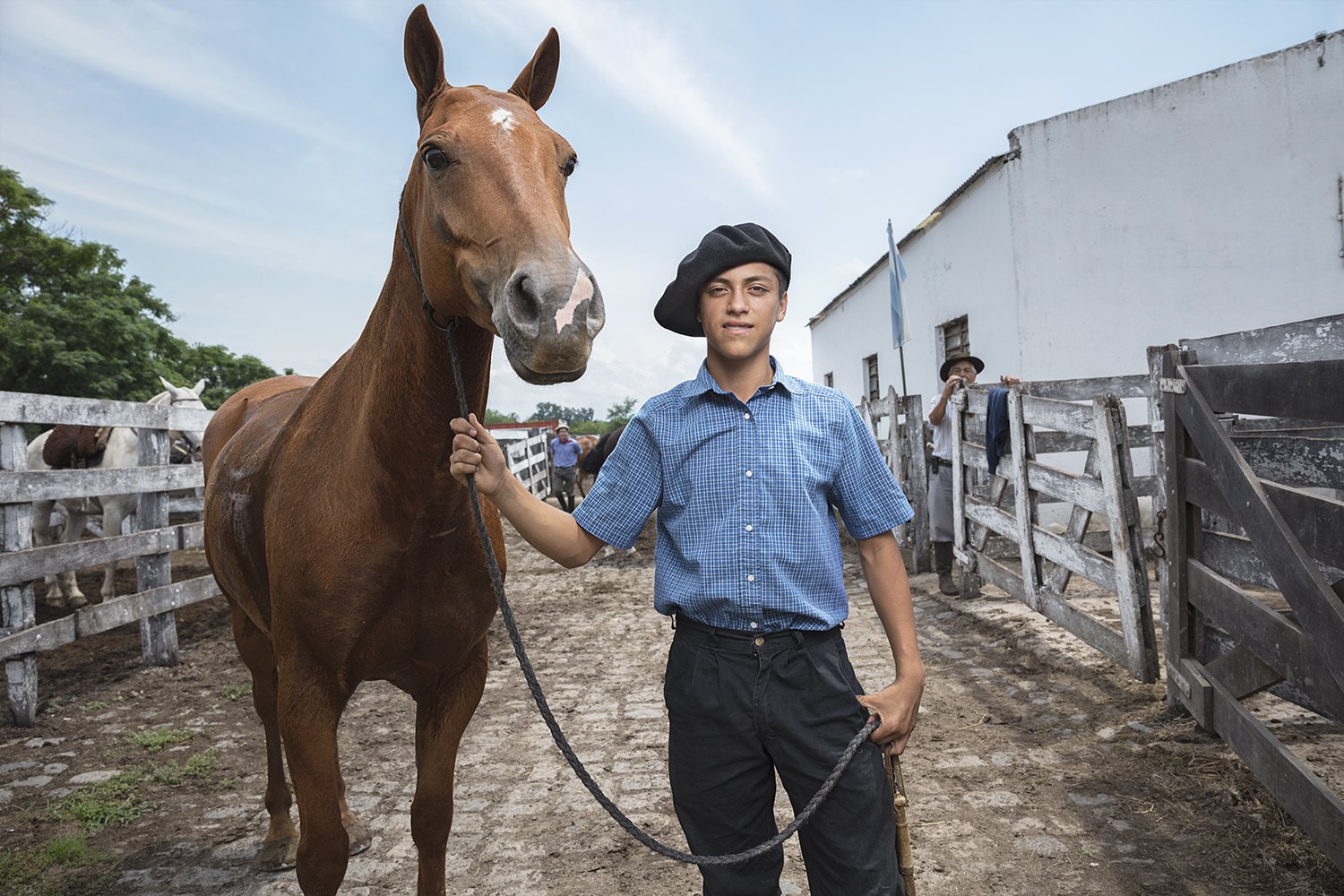 A young man in a boina leads a chestnut horse down the alley between the corrals
