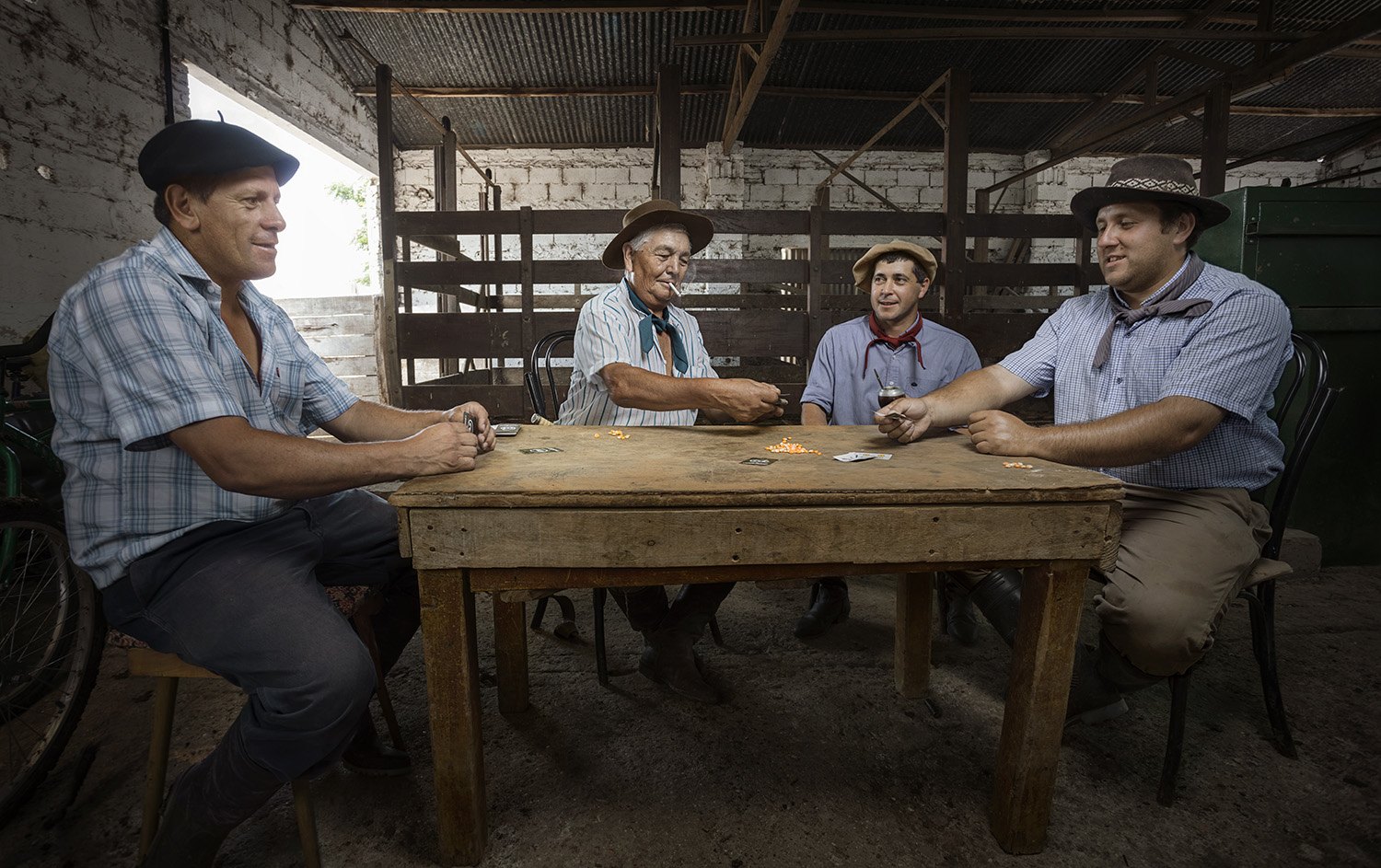 Four men playing cards at a wooden table in the shade of an open barn