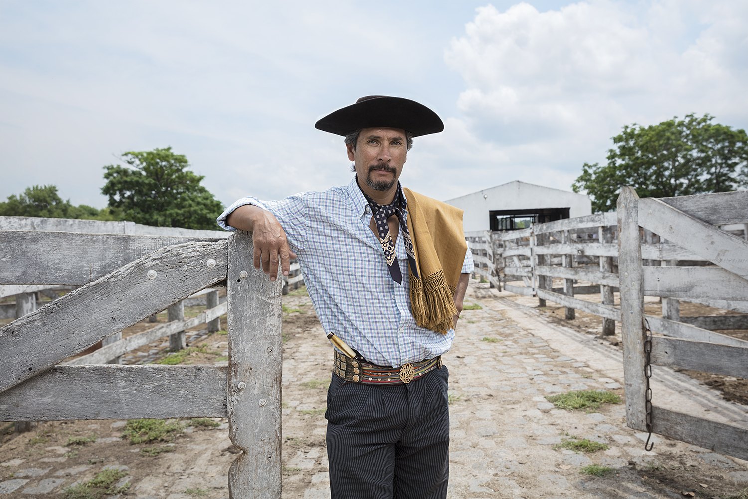 A gaucho in a flat-brimmed black hat rests an arm on a fence post, silver belt catching the light