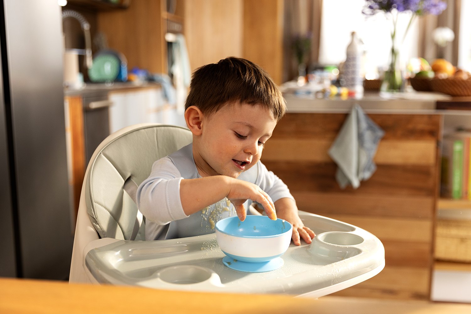 A toddler in a high chair reaches into a small blue bowl, warm kitchen out of focus behind