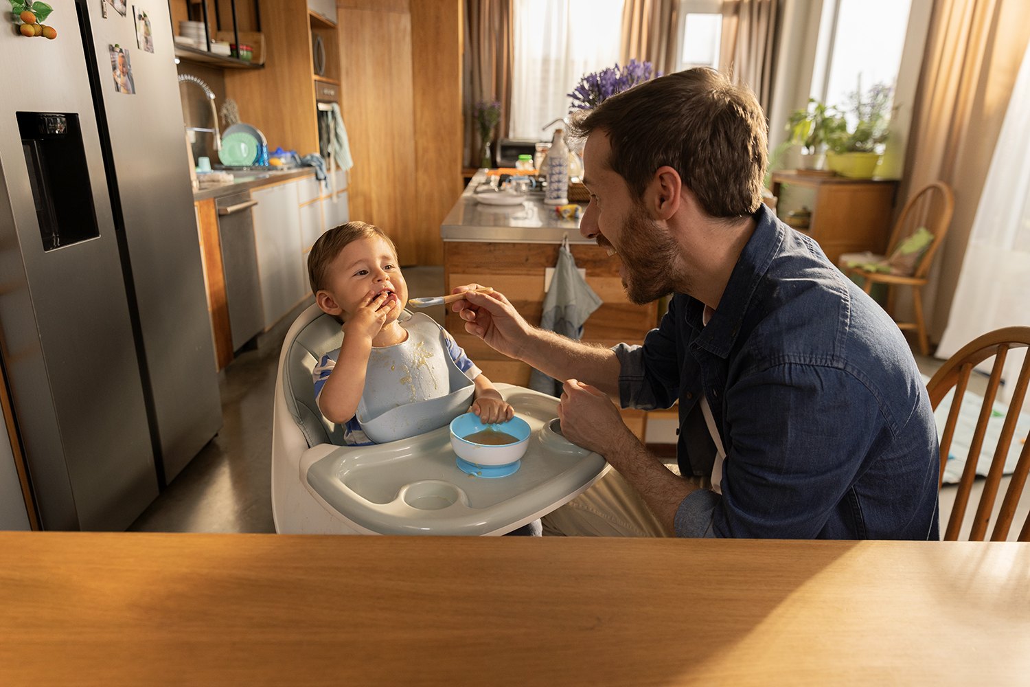 A father in a denim shirt leans in with a spoon while his son eats in the high chair