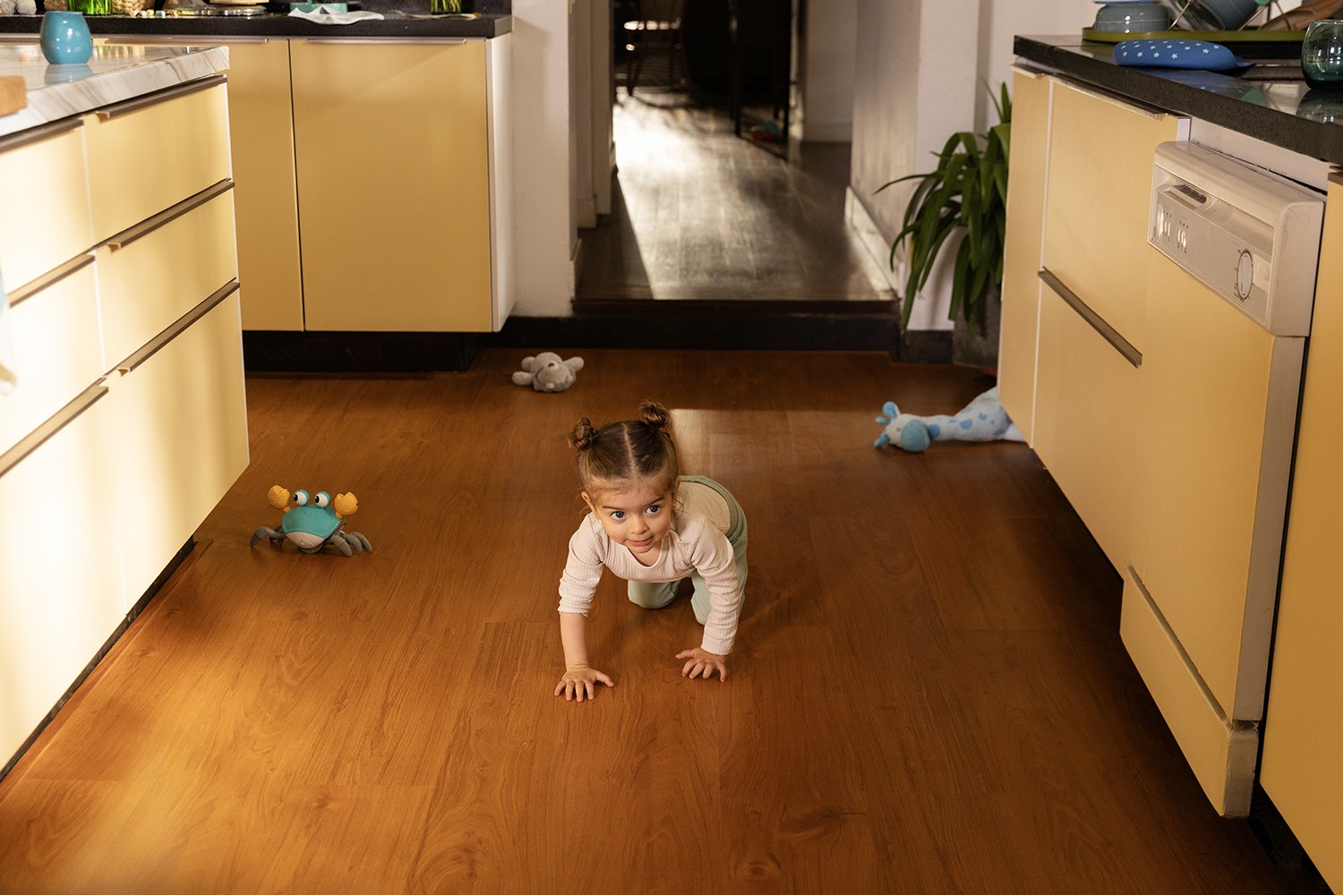 A little girl in pale clothes crawls down the wooden kitchen floor towards the camera