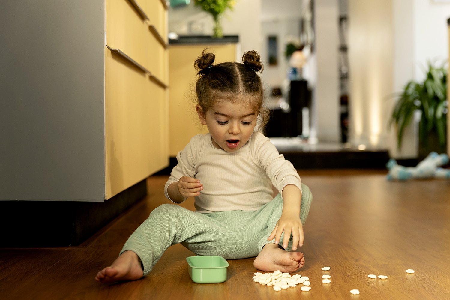Sitting on the floor beside a green bowl with cereal scattered in an arc around her