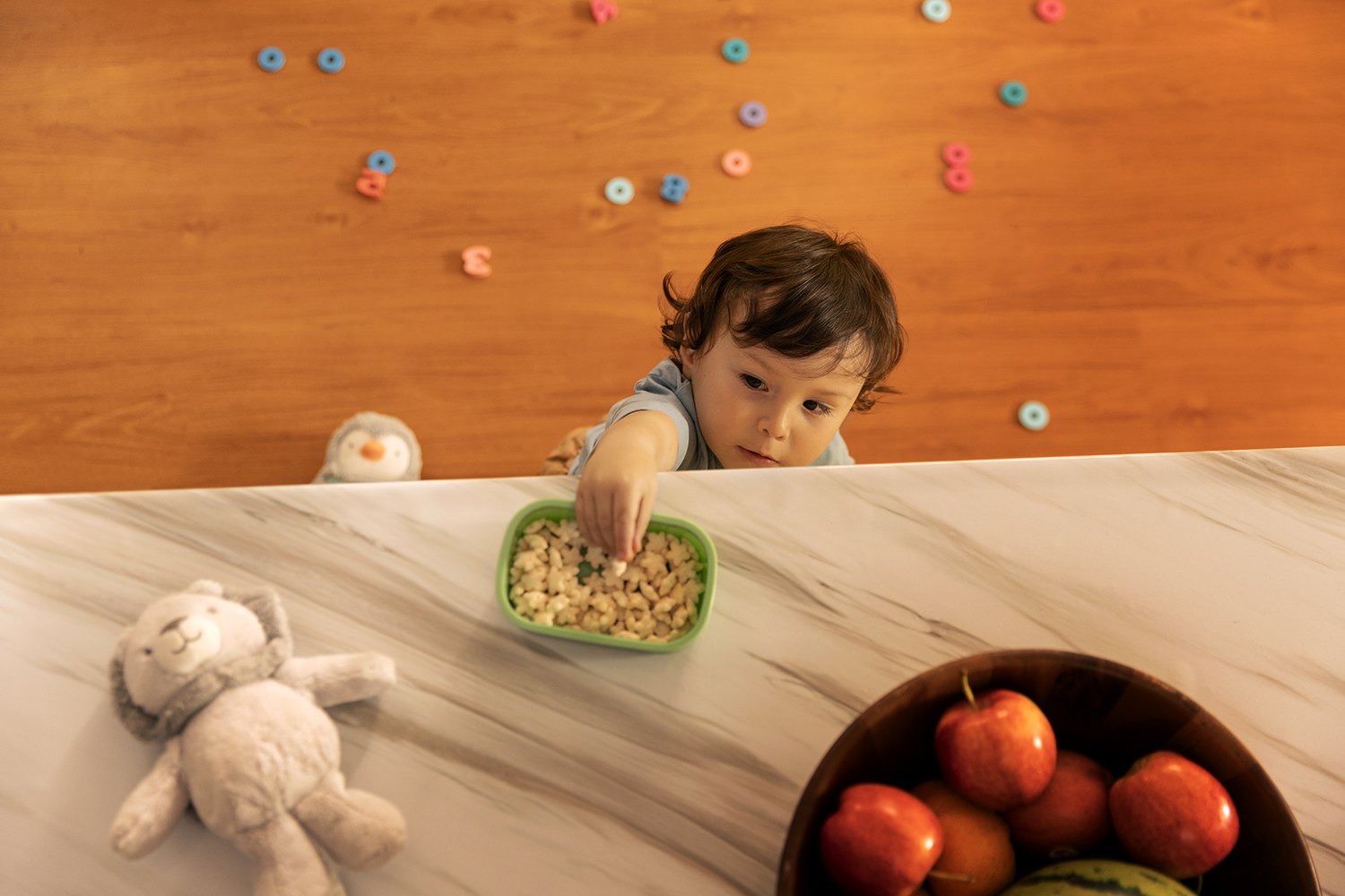 A child reaches across the marble counter for a green bowl, cereal thrown across the wood behind
