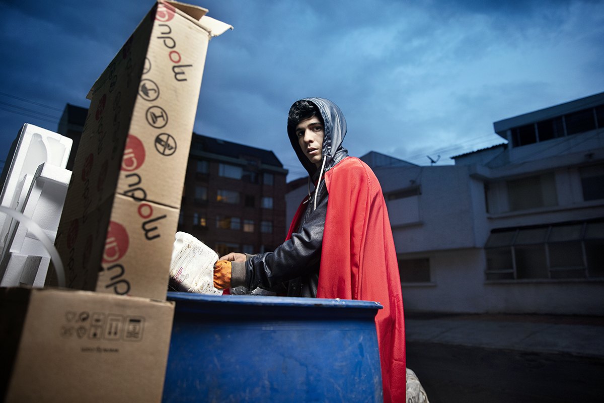 A hooded recycler in a red cape leans on a blue wheelie bin against a stormy night sky