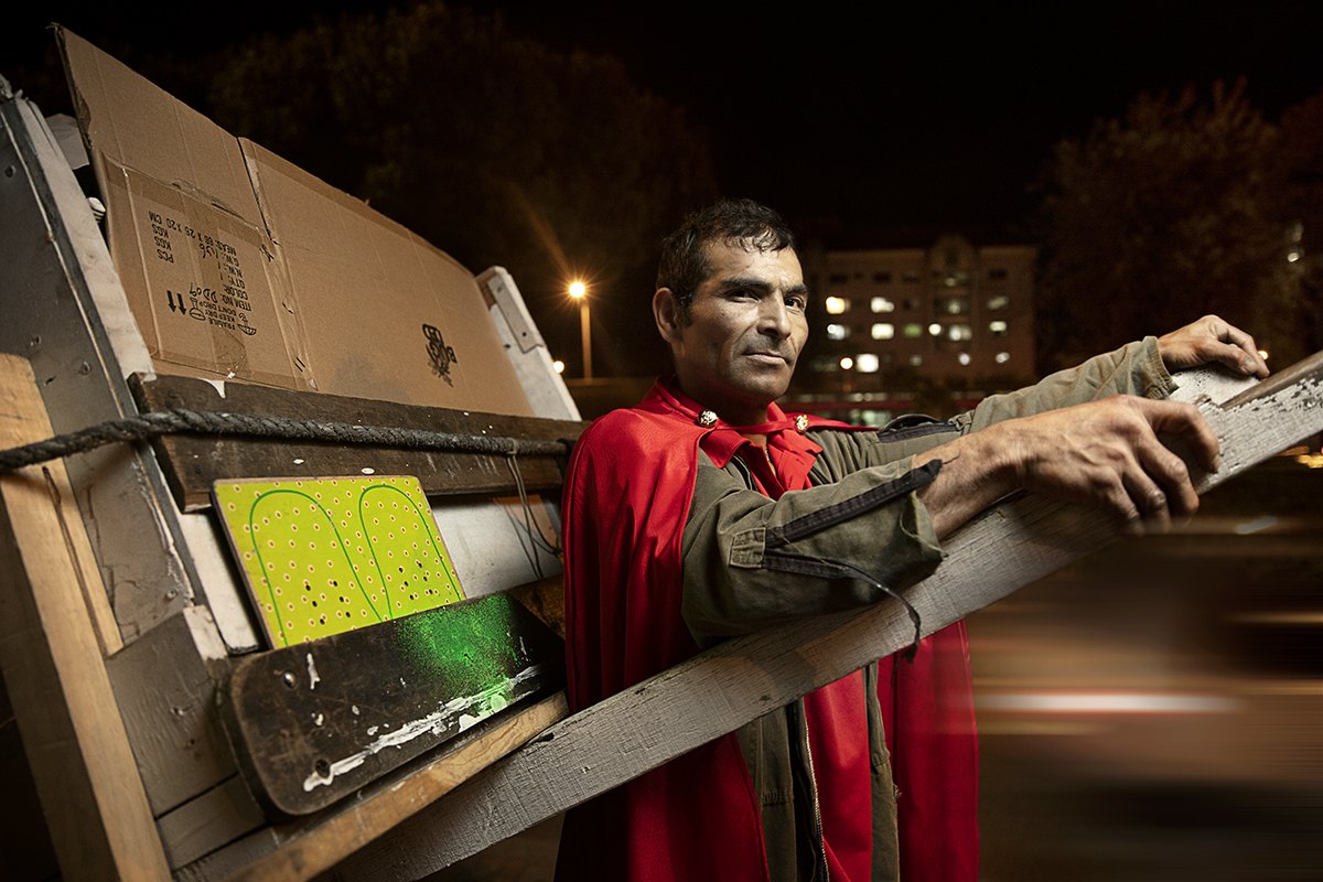A caped recycler gripping the frame of his loaded cart, city lights behind