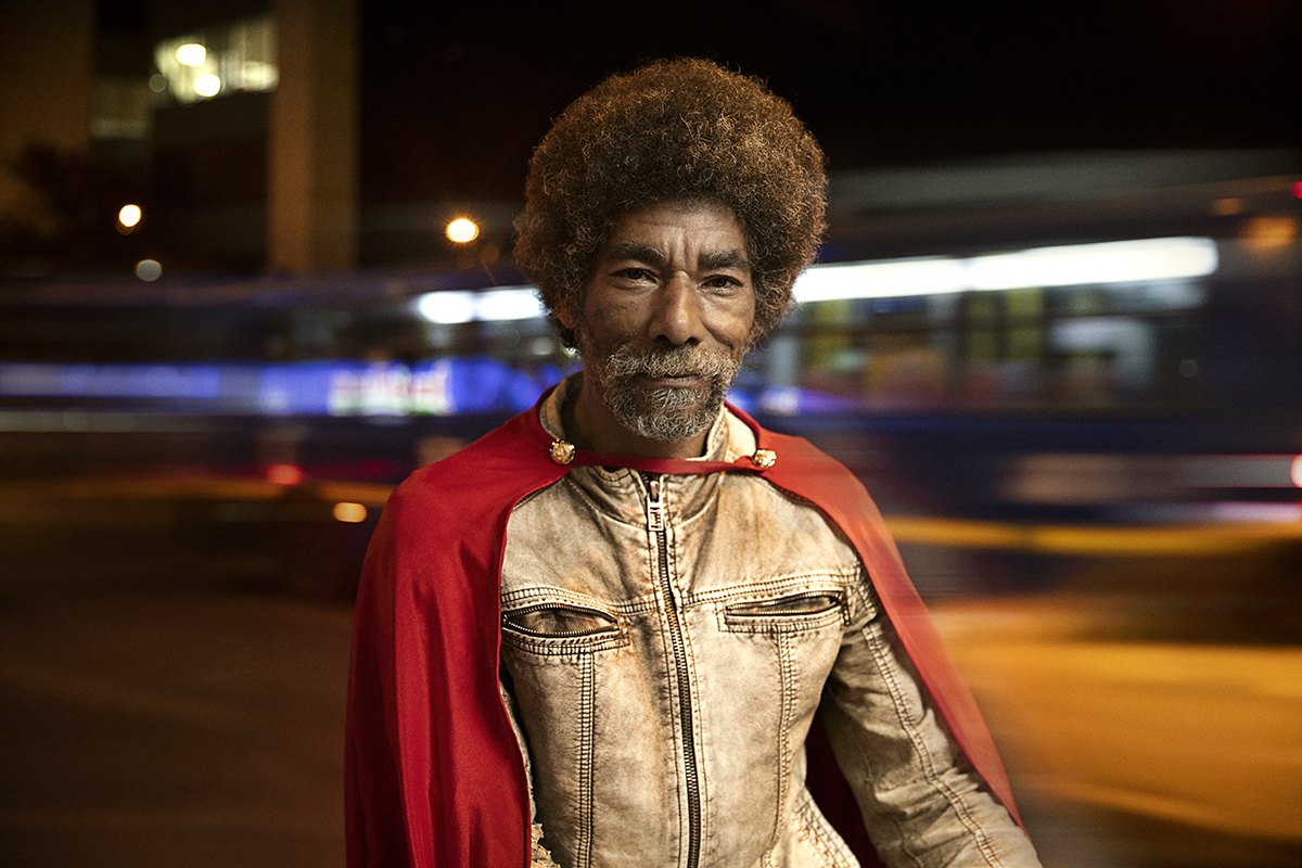 Portrait of an older recycler in a red cape, traffic streaking past behind him