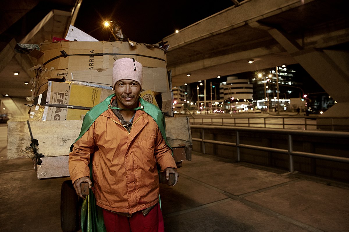 A recycler in an orange jacket in front of a cart piled twice his height with cardboard