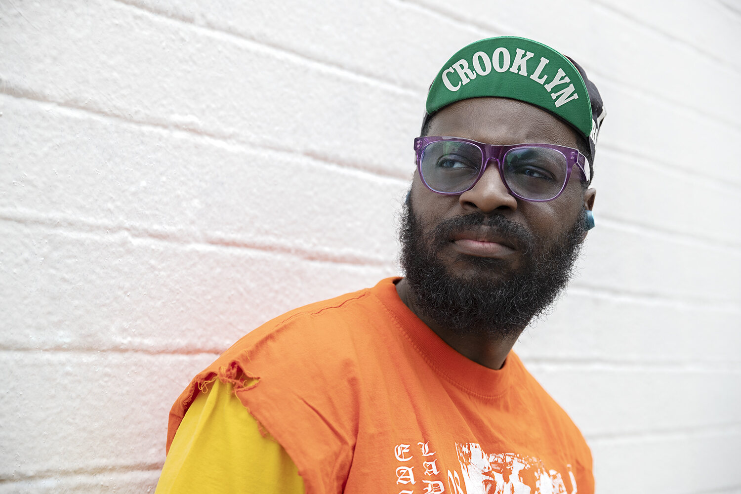 Orange tee and green Crooklyn cap against a white brick wall