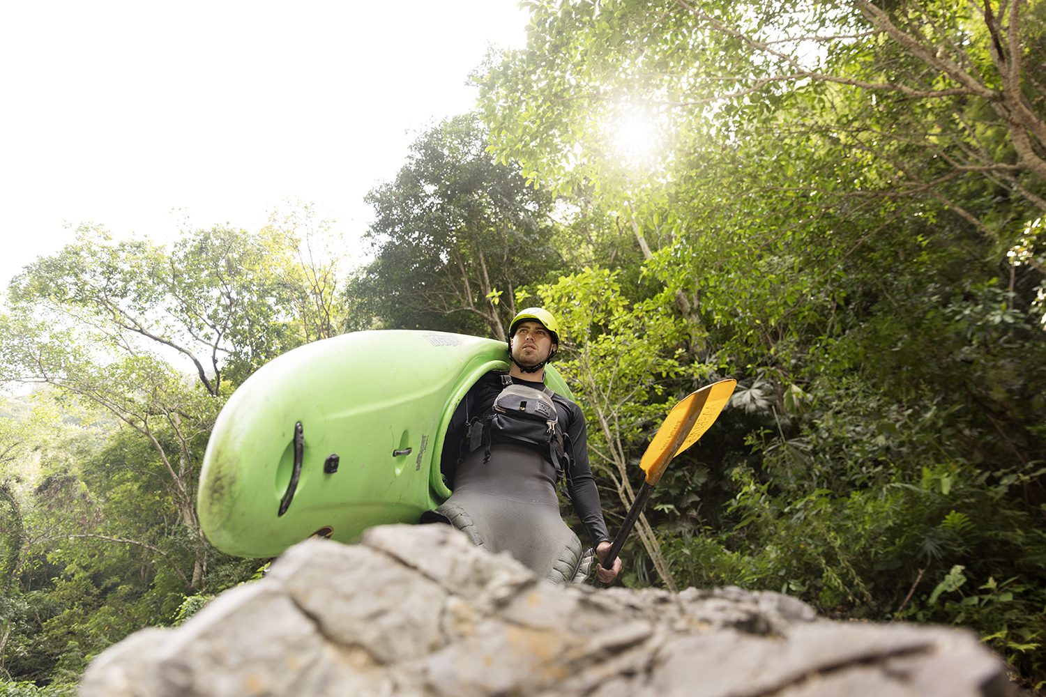 A kayaker rests on a rock with the boat, sun through the jungle